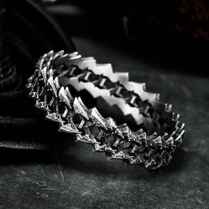 Op de afbeelding: Een zilverkleurige armband met een gedetailleerd, gestructureerd ontwerp. De armband heeft een reeks onderling verbonden, puntige elementen, waardoor een robuuste, industriële esthetiek ontstaat. Het sieraad wordt op een donkere ondergrond gepresenteerd.