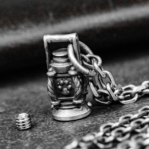 May include: A silver pendant necklace with a miniature lantern design. The lantern has detailed features, including a paw print emblem, and is connected to a chain. A small, threaded component is also visible.