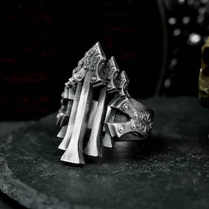 Puede incluir: Un anillo plateado detallado con un diseño intrincado. El anillo presenta una serie de elementos verticales en forma de cuchilla y detalles ornamentados en la banda. La estética general es gótica y atrevida.