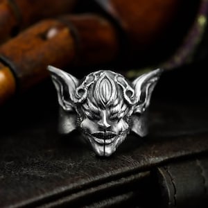 Puede incluir: Un anillo plateado con un diseño detallado de una cara estilizada, con orejas grandes y una boca cosida. El intrincado diseño del anillo se destaca sobre un fondo oscuro.