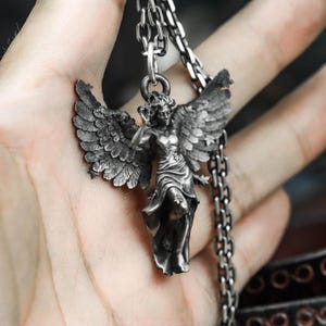 May include: A silver-toned pendant necklace featuring a detailed angel figure with outstretched wings. The angel is depicted in a flowing gown, with intricate feather detailing on the wings. The necklace has a chain with a lobster clasp.