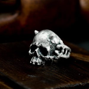 May include: A silver-colored skull ring with small horns and a cracked surface. The ring has a detailed skull design with eye sockets and teeth. The ring is set against a dark wooden background.