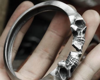 May include: A silver-colored bracelet with a skull design. The bracelet features two skull heads facing each other, with a broken section between them. The bracelet is held in a hand.