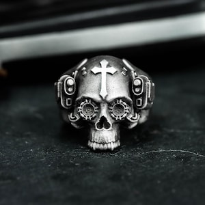 May include: A silver skull ring with a cross on the forehead and intricate mechanical details around the eyes and sides. The ring has a gothic or biker theme, with a detailed, textured surface.