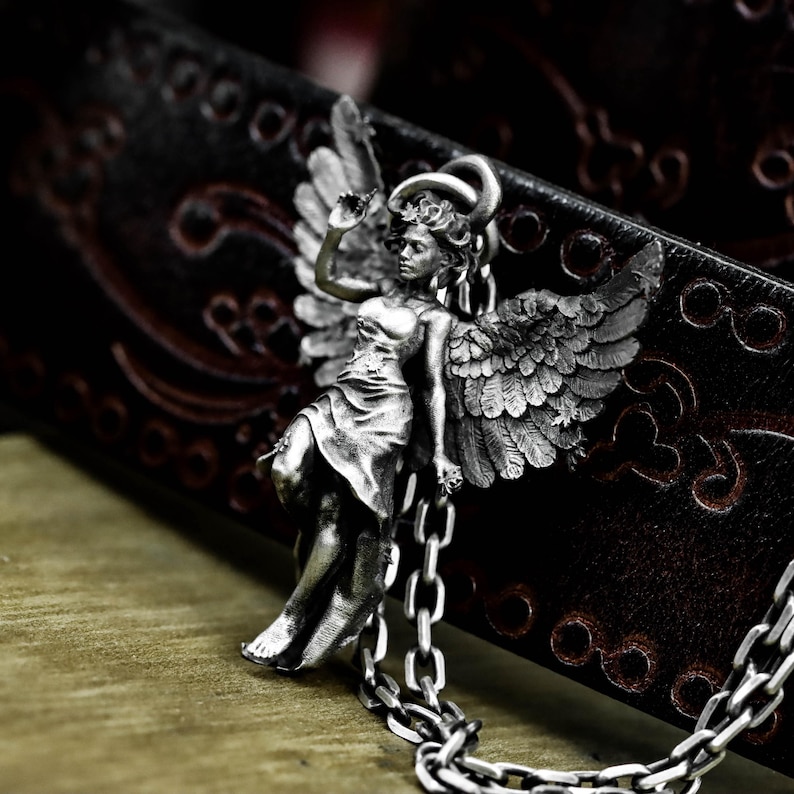 May include: A silver-toned pendant necklace featuring a detailed angel figure with outstretched wings. The angel is depicted in a flowing gown, with a halo and chain. The necklace is set against a dark, textured background.