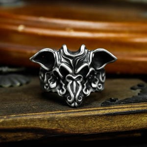 May include: A detailed silver-colored ring with an ornate, gothic design. The ring features a stylized creature with prominent ears and intricate patterns. The ring is displayed on a wooden surface, showcasing its detailed craftsmanship.