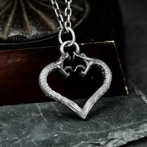 May include: A silver-toned pendant necklace featuring a heart-shaped design. The heart is formed by two detailed snake heads facing each other. The snakes' bodies create the heart shape, and the necklace hangs from a silver chain.