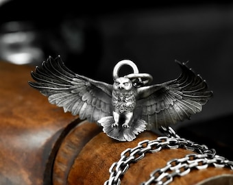 May include: A detailed silver owl pendant with outstretched wings, attached to a silver chain. The owl has intricate feather detailing and a small loop at the top for the chain. The pendant is displayed on a wooden surface.