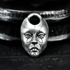 Op de afbeelding: Een zilverkleurige hanger in de vorm van een gestileerd gezichtsmasker. Het masker heeft gedetailleerde lijnen en een lus aan de bovenkant voor een ketting. Het ontwerp heeft een futuristische, robotachtige uitstraling, met focus op de gelaatstrekken.