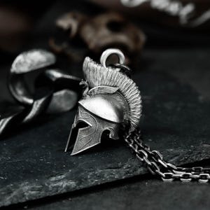 Puede incluir: Un collar con colgante plateado que presenta un casco espartano en miniatura detallado. El casco tiene una superficie texturizada, una cresta prominente y una cadena. El colgante se encuentra sobre un fondo oscuro y texturizado.