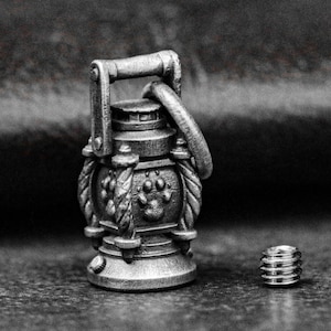 May include: A detailed, monochrome miniature lantern with a handle and paw print design. A small, threaded component sits beside it. The design suggests a vintage or fantasy theme.