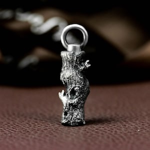 Könnte beinhalten: Ein silberfarbener Anhänger in Form eines Baumstamms. Der Anhänger hat eine strukturierte Oberfläche mit einem Ring oben zum Befestigen an einer Kette. Das Design umfasst Details wie Rinde und kleine Äste.