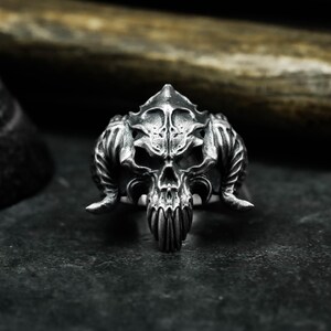 May include: A detailed silver-colored ring featuring a skull design with prominent horns and intricate facial details. The ring is set against a dark, textured surface, highlighting the gothic and edgy aesthetic of the jewelry.