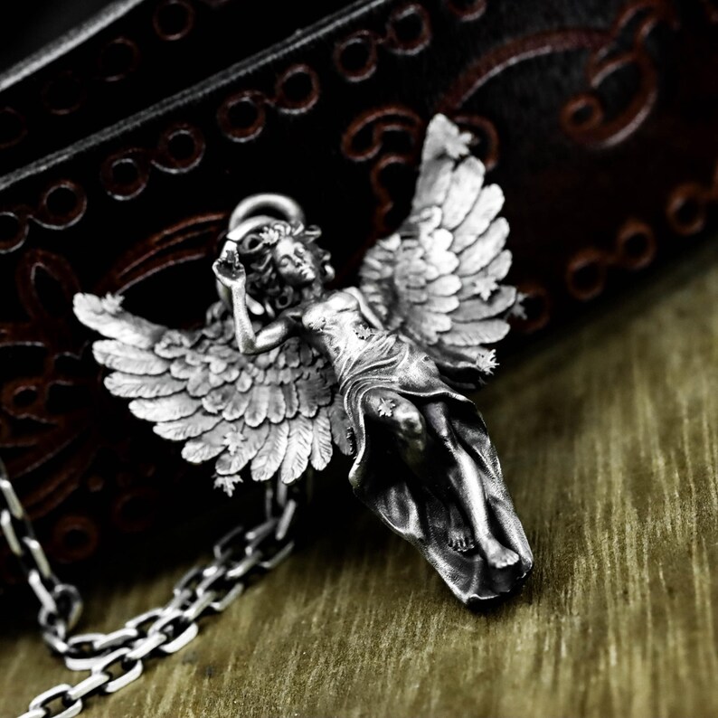 May include: A silver-colored angel pendant with outstretched wings and a halo. The detailed pendant is attached to a silver chain. The angel is lying down with one arm raised. The background is a dark brown leather-like surface.