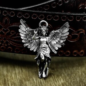 May include: A silver-colored angel pendant with detailed wings and a flowing dress. The pendant is set against a dark brown leather belt with decorative stitching, creating a contrast of textures and tones. The angel is in a graceful pose.