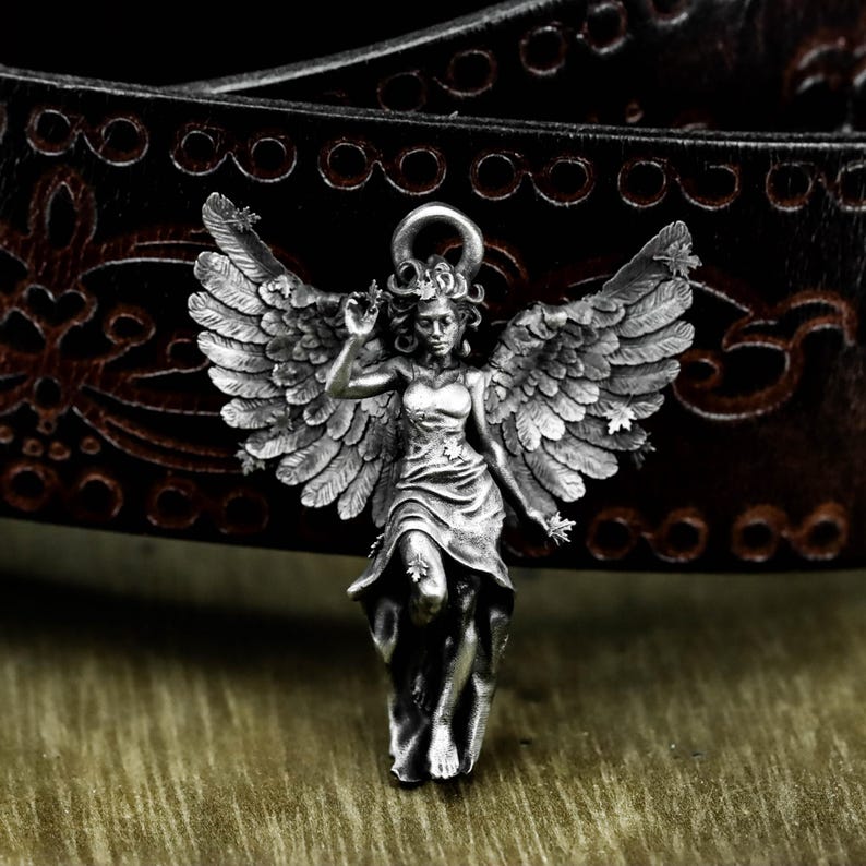 May include: A silver-colored angel pendant with detailed wings and a flowing dress. The pendant is set against a dark brown leather belt with decorative stitching, creating a contrast of textures and tones. The angel is in a graceful pose.