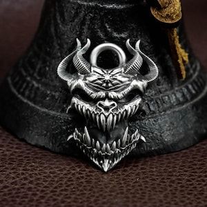 May include: A detailed silver pendant in the shape of a demonic face with sharp teeth and horns. The pendant has a textured surface and a loop at the top for a chain. The background is dark, highlighting the intricate design of the jewelry.