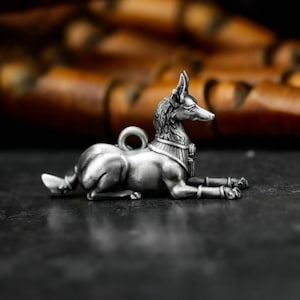 Może przedstawiać: Srebrny wisiorek Anubisa w pozycji spoczynku. Wisiorek przedstawia boga z głową szakala ze szczegółowymi cechami, w tym kołnierzem i pętlą do zawieszenia. Tło jest rozmyte, z ciemną powierzchnią.