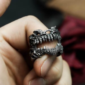 May include: A silver-colored ring with a detailed design of a dragon's open jaws. The ring features sharp teeth and textured scales, creating a bold and gothic aesthetic. The ring is worn on a finger, showcasing its intricate craftsmanship.
