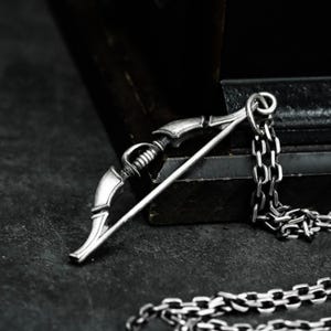 Puede incluir: Un collar con colgante plateado que representa un arco y una flecha. El colgante, con detalles finos, cuelga de una cadena a juego. El arco presenta detalles intrincados y la flecha es recta. El collar se presenta sobre un fondo oscuro.