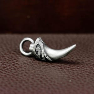 May include: A silver-colored pendant in the shape of a curved horn or claw. The pendant has a loop for a chain and decorative swirls on the side. The horn is smooth and polished, with a sharp point.