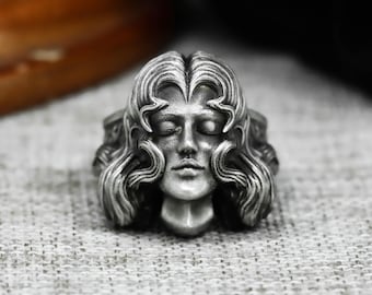 Könnte beinhalten: Silberner Ring mit einer detaillierten Skulptur eines Frauengesichts mit geschlossenen Augen und fließendem Haar. Der Ring hat eine Vintage-Ästhetik mit aufwendigen Details an Gesicht und Haar.