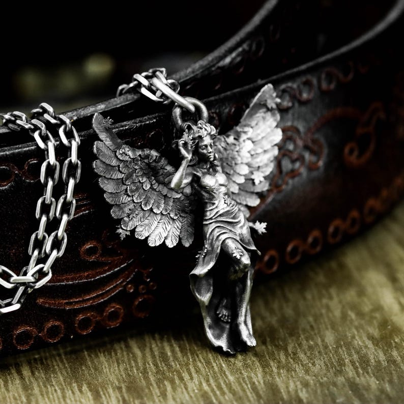 May include: A silver-colored pendant necklace featuring a detailed angel figure with outstretched wings. The angel is depicted in a flowing gown, with intricate feather detailing on the wings. The necklace chain is a chunky, silver-toned link. The background is a dark brown leather.