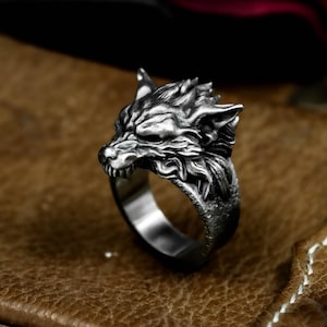 May include: A silver-colored ring featuring a detailed wolf head design. The wolf's face is intricately sculpted, showcasing sharp teeth and textured fur. The ring has a polished band and is set against a brown leather surface.