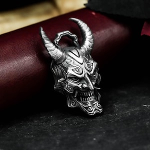 Può includere: Un ciondolo argentato dettagliato a forma di maschera Oni, con corna ricurve, denti affilati e dettagli del viso intricati. Il ciondolo ha un anello nella parte superiore per una catena ed è posto su uno sfondo scuro.