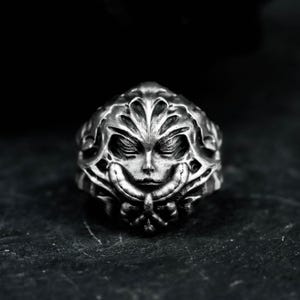 May include: A silver-toned ring featuring a detailed face design with closed eyes and ornate floral or leaf-like embellishments. The ring has a textured appearance and is set against a dark background.