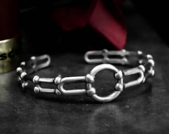 Könnte beinhalten: Ein silberfarbenes Armband mit einem einzigartigen Design. Das Armband hat ein zentrales kreisförmiges Element, das durch U-förmige Glieder und kleinere dekorative Details verbunden ist. Das Metall hat ein leicht gealtertes Aussehen.