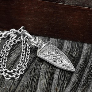 May include: A silver-colored pendant necklace featuring a stylized arrowhead design. The pendant has intricate engraved patterns and is suspended from a matching chain. The necklace is displayed on a textured surface.