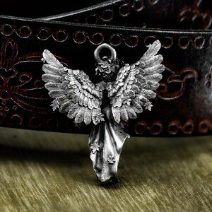 May include: A detailed silver angel pendant with outstretched wings. The pendant features intricate feather and leaf details, with a loop at the top for a chain. The background shows a dark brown leather belt with embossed patterns.