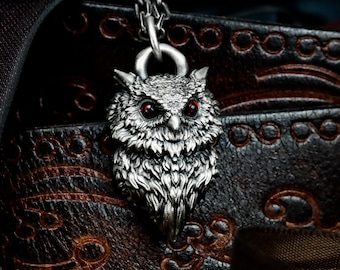 May include: A silver owl pendant with red eyes hangs from a chain. The owl has detailed feathering and a stern expression. The pendant is set against a dark, textured background, creating a striking contrast. The owl's eyes are a deep red.