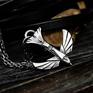 Può includere: Collana con pendente color argento raffigurante un uccello stilizzato in volo. L'uccello ha ali e piume della coda dettagliate, sospese a una catena. La collana è presentata su una superficie di legno scuro, con uno sfondo sfocato.