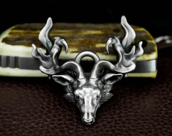 May include: A silver-colored pendant featuring a detailed ram's head with large, curved horns. The pendant has a loop at the top for a chain and is set against a brown leather surface. The ram's head is the focal point, showcasing intricate details.