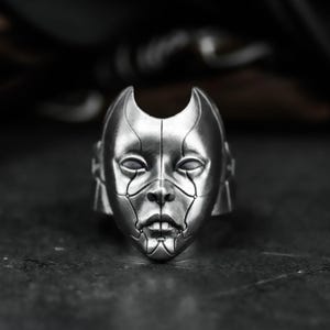 Op de afbeelding: Een zilverkleurige ring met een gestileerd gezichtsontwerp. Het gezicht heeft een futuristische, robotachtige uitstraling met vloeiende lijnen en een symmetrische indeling. Het ontwerp van de ring omvat een prominente neus, lippen en ogen, met gebogen elementen aan de bovenkant.