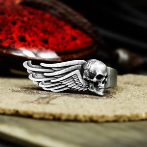 Peut inclure: Bague argentée ornée d'un crâne détaillé avec des ailes. Le crâne présente des traits précis, et l'aile est finement travaillée. La bague est posée sur une surface texturée. Un bijou.