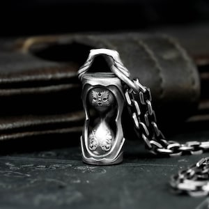 Może przedstawiać: Srebrny wisiorek w kształcie klepsydry z misternymi detalami, zawieszony na pasującym łańcuszku. Wisiorek posiada ozdobne wzory i kaptur, tworząc gotycką lub fantastyczną estetykę. Łańcuszek jest widoczny.