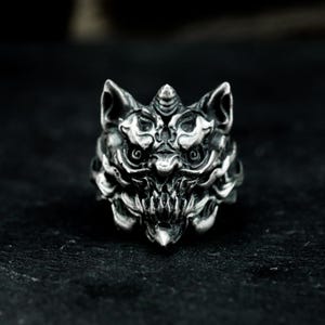 May include: A detailed silver ring depicting a stylized, fierce creature's face. The ring displays intricate detailing of the creature's eyes, teeth, and facial features. The ring is a bold statement piece, ideal for those who appreciate unique jewellery.