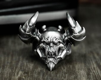 May include: A silver-colored ring featuring a detailed, demonic skull design. The ring has prominent horns and intricate facial features, creating a gothic or fantasy aesthetic. The ring is likely made of metal.