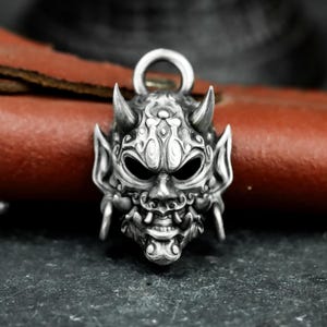 May include: A detailed silver pendant in the shape of a Japanese Oni mask. The pendant features intricate carvings, horns, and a loop for a chain. The mask has a fierce expression with visible teeth and large eyes, showcasing a detailed design.