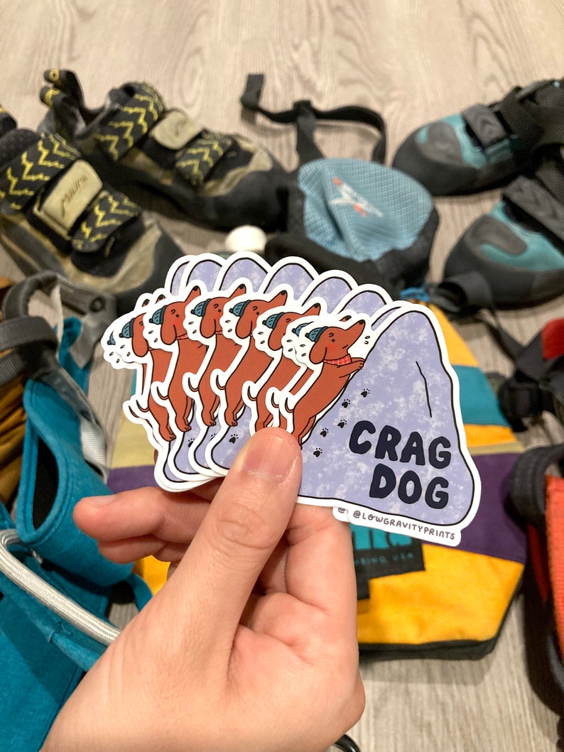 Hand holding a stack of stickers, crag dog, climber life, cragdogs, dog lovers, adorable sticker of wiener dog, climbing shoes, chalk bag, carabiner, harness, climbing rope, belay card, toprope, on belay, r/climbing, r/climbergirls, sloper rock