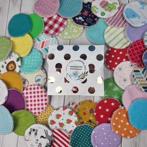 Könnte beinhalten: Eine Sammlung bunter wiederverwendbarer Abschminkpads mit verschiedenen Mustern, darunter Punkte, Streifen und Blumenmuster. Die Pads sind in einem Kreis um einen kleinen weißen Umschlag mit dem Text "Wundertiite Abschminkpads Bunt" angeordnet.