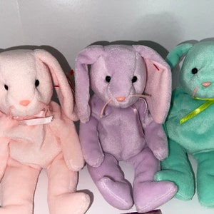 May include: Three pastel-coloured plush bunny toys: pink, lavender, and mint green. Each bunny has long ears, a pink nose, and a ribbon or bow. The bunnies are soft and cuddly, ideal for Easter or as a gift.