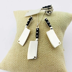 May include: A set of three silver cleaver-shaped pendants with black handles. The pendants are displayed on a beige fabric surface. The largest pendant is a necklace, the medium pendant is a brooch, and the smallest pendant is an earring.