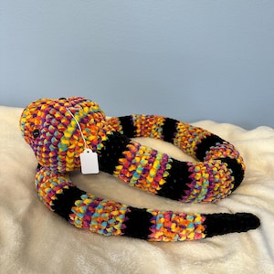 May include: A handmade, plush snake toy with a vibrant, multicolored head and body. The body features alternating black and rainbow-colored stripes. The snake is lying on a soft, white surface.