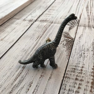 May include: A decorative dinosaur-shaped cabinet knob in a distressed, dark gray finish. The knob features a long neck and tail, with a bolt for easy installation. The dinosaur is approximately 4 inches tall.