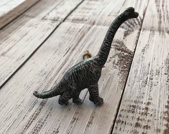 May include: A decorative dinosaur-shaped cabinet knob in a distressed, dark gray finish. The knob features a long neck and tail, with a bolt for easy installation. The dinosaur is approximately 4 inches tall.