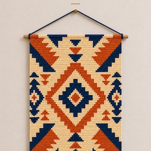 Op de afbeelding: Een gehaakte wandhanger met de tekst SOUTHWEST GEO CROCHET WALL HANGING PATTERN. Het rechthoekige textiel toont een geometrisch patroon in crème, gebrand oranje en marineblauw, opgehangen aan een houten stok met een marineblauw koord.
